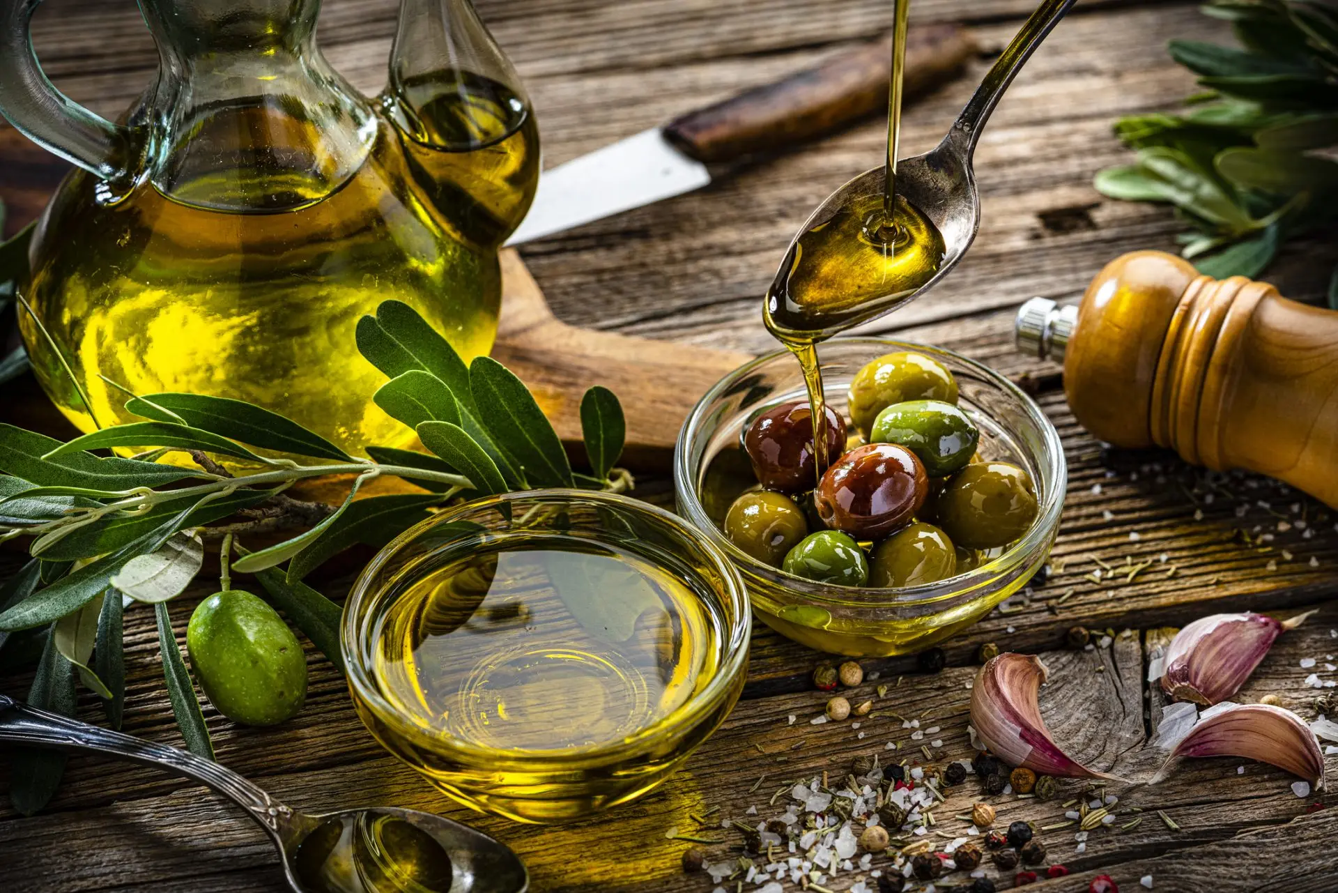 یک ظرف بزرگ روغن زیتون با برگه‌های زیتون و یک قاشق چوبی در کنار آن